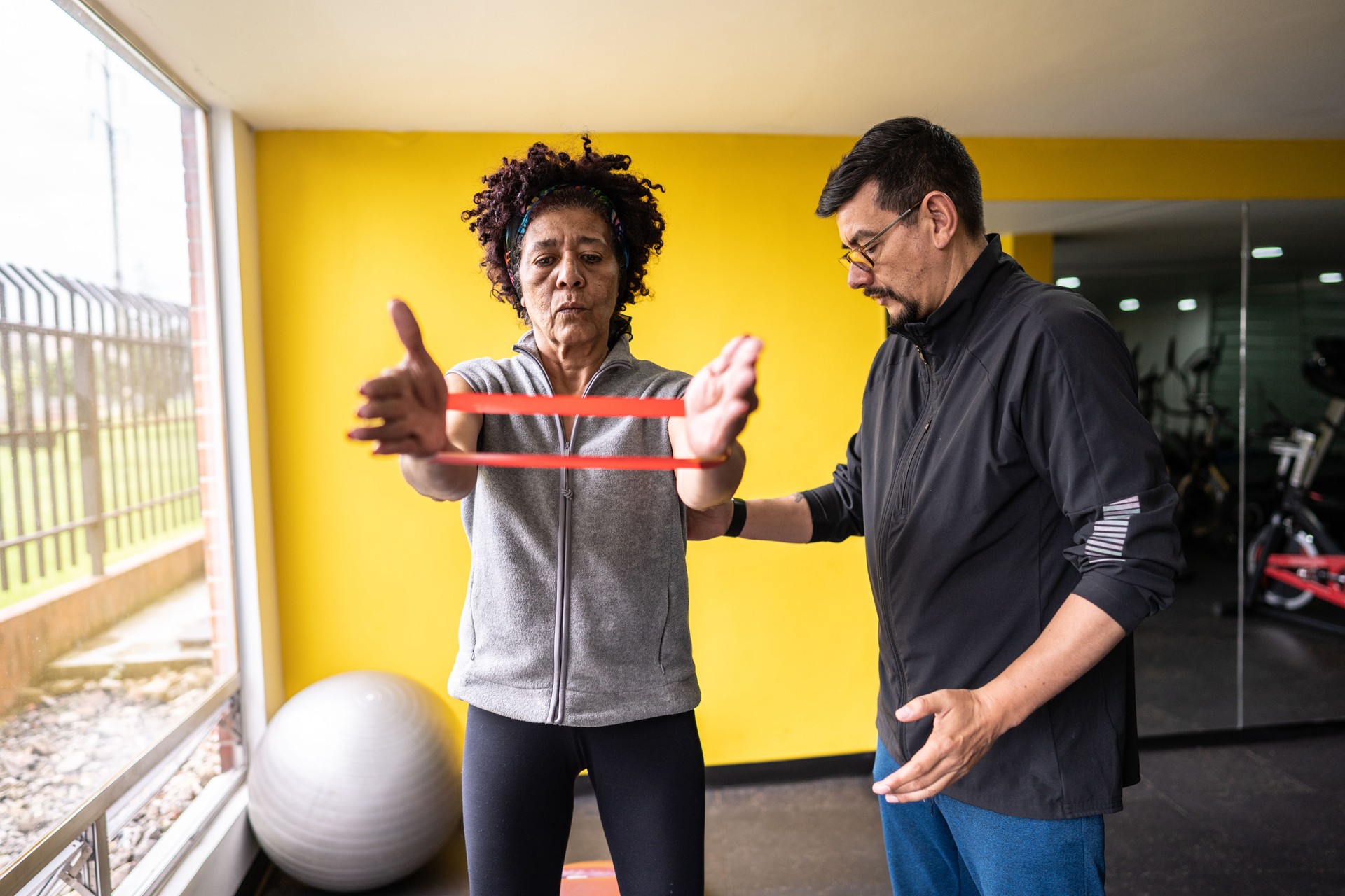 Senior woman exercising using a resistance band with assistance from fitness instructor at gym